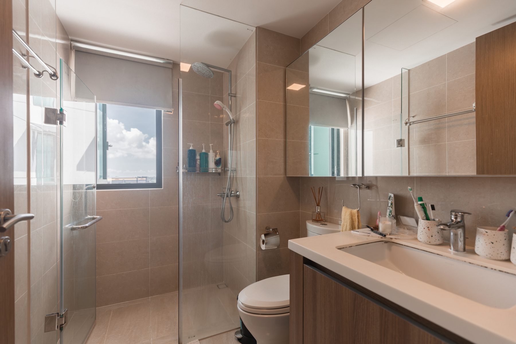 Modern bathroom with glass-enclosed shower, beige tiles, vanity, and window for natural light