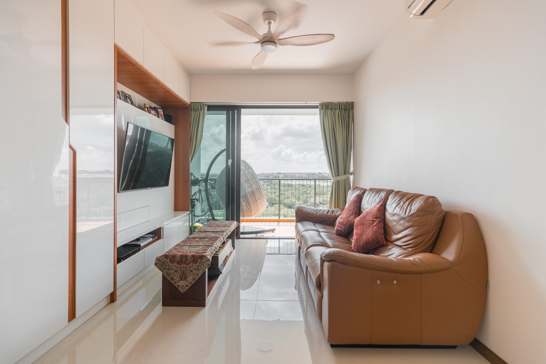 Living room with brown leather sofa, ceiling fan, TV unit, and balcony with forest view